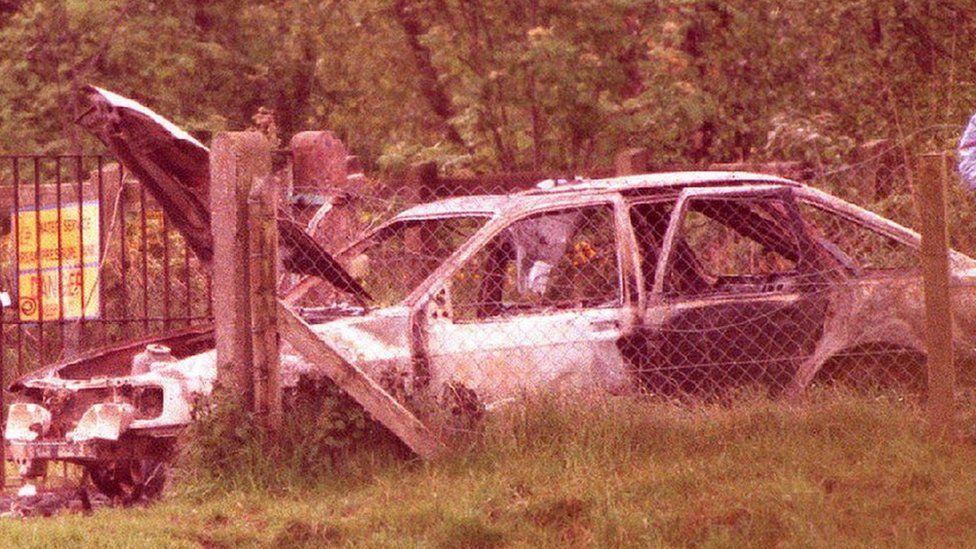 Old image of a burnt out white car behind a wired fence