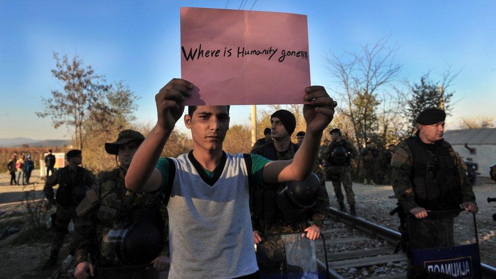 A migrant holds up a sign reading "Where is humanity gone" as he is surrounded by security