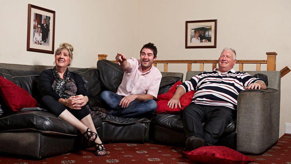 A promotional photo of George Gilbey on a sofa, pointing away from the camera and laughing, wearing a pink shirt. His mother Linda and stepfather Pete are sat either side of him.