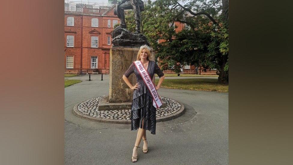 A woman is standing in front of a statue, wearing a dress with a sash that reads "miss Shropshire 2025". She has blonde hair and a silver tiara.