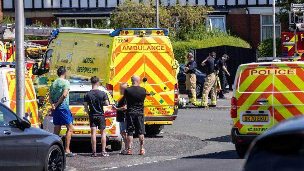 A range of emergency vehicles are parked up. Emergency service workers stand around. Properties can be seen in the background.