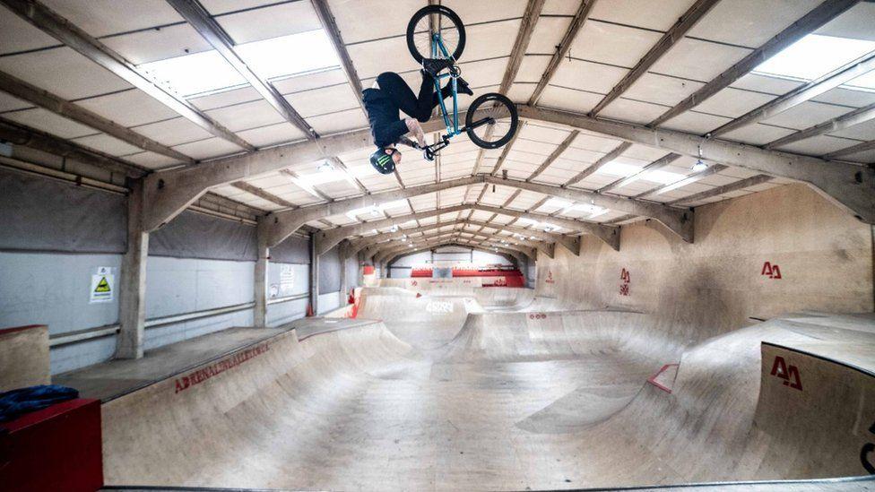 Wooden indoor BMX quarter pipe extending throughout a long building.  A BMX bike and rider are seen upside down on the ceiling.