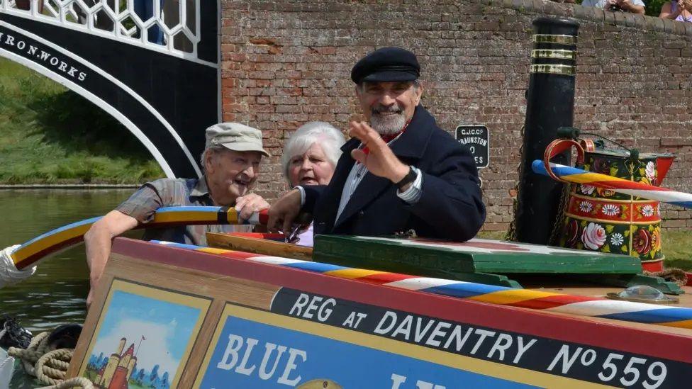 Sir David Suchet with dark hair and beard wearing a dark-coloured cap and rdark-coloured jacket is at the stern of a narrowboat moving the tilled with one hand and waving with the other. There is a man in a light brown cap and a woman with white hair behind him. There is a painted water can on the roof of the boat alongside a chimney. The boat has colourful paintwork and the words REG AT DAVENTRY in white lettering on the side. The boat has just passed through an iron bridge painted in black and white.