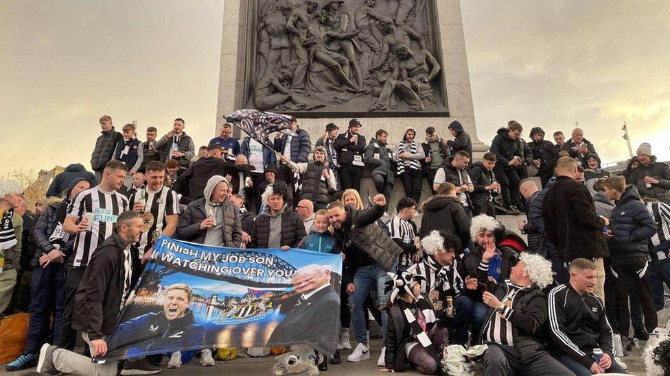 A large group of people, many in Newcastle United shirts, stand at the base of Nelson's Column