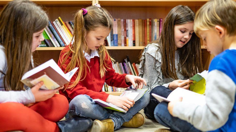 A group of children reading in a circle. Three girls and a boy are reading books with a book shelf behind them.