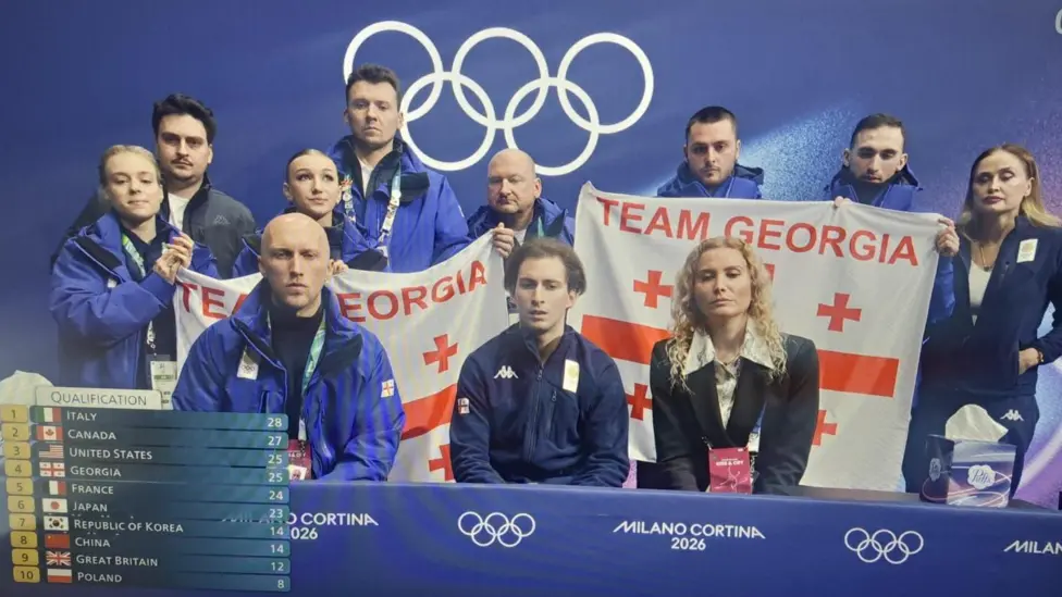 The Georgia team gather together to learn their scores, holding up two Team Georgia flags and showing serious facial expressions
