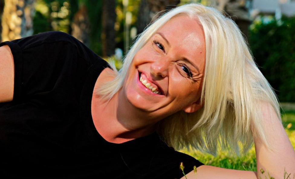 A photo of Hollie laying on the grass outside smiling with long blonde hair before her cancer diagnosis