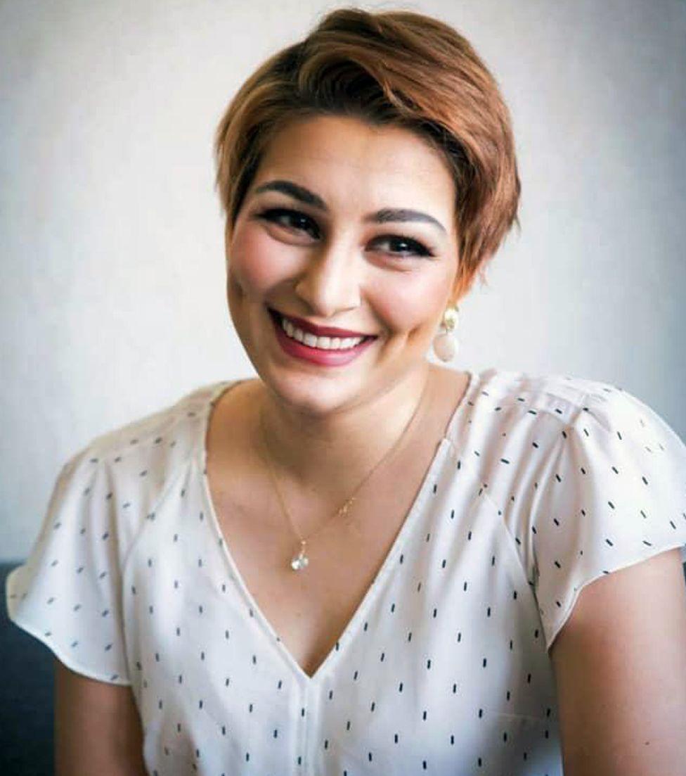 A photograph of Vida against a light grey background. Vida is smiling and has short brown hair. She is wearing make-up, a thin gold chain around her neck and a short-sleeved V-necked top which is white with small dark spots. 