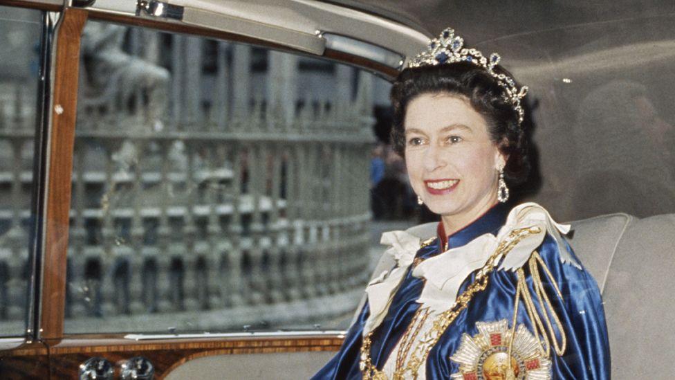 Queen Elizabeth, in royal regalia including a crown, smiling and riding in a car in the late 1960s.