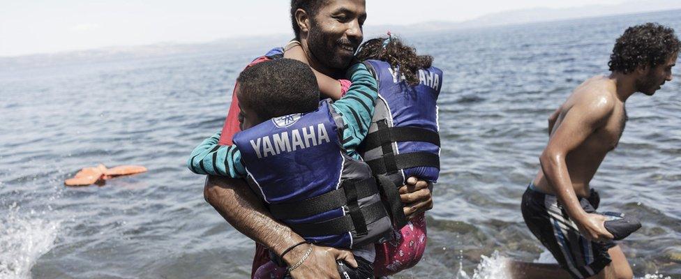 A smiling migrant carries his two children as he gets off an inflatable boat after arriving on the Greek island of Lesbos after crossing from Turkey to Greece on 14 August 2015