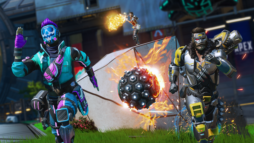 Two characters in futuristic armour sprint towards the viewer. The character on the left wears a sky blue suit of armour with a skull-face mask and bright purple trim. The character on the right wears a silver suit of armour with yellow trim and a mask styled on a gorilla's face. They're fleeing from a large, spiked metal ball with flames emanating from it. Behind them, stood atop a piece of wreckage, a third character fires at an unseen enemy - a volley of flame emanating from their weapon.