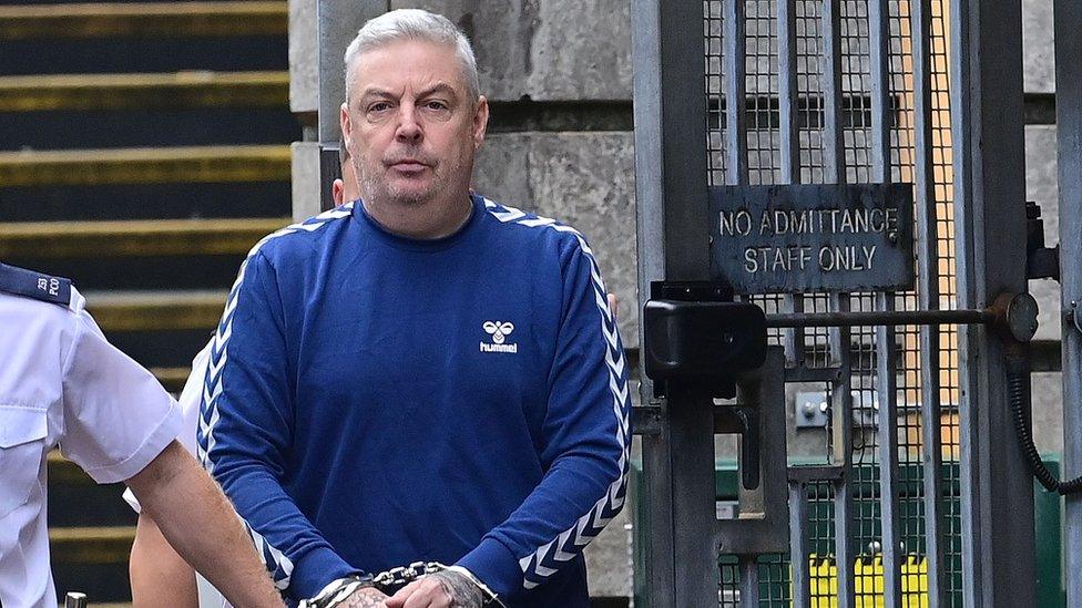 Christopher O'Kane, wearing a blue sweater, looks at the camera as he is led into court in handcuffs