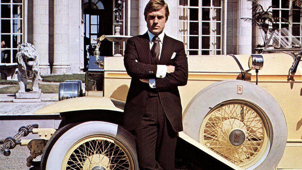 Robert Redford is seen in a still photo promoting The Great Gatsby. He is standing in front of a vintage yellow Rolls Royce and has his arms crossed
