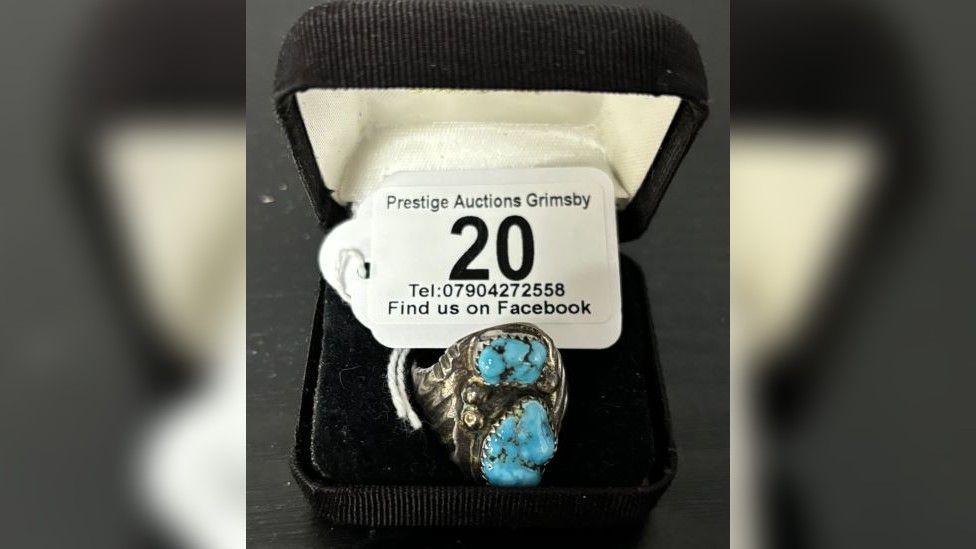 A silver ring in a black ring box with a white label on it stating "Prestige Auctions Grimsby". The ring is silver with indentations and turquoise stones. 