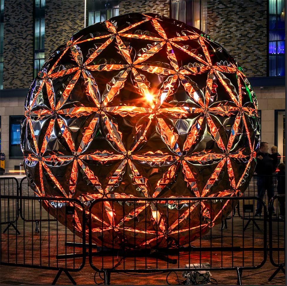 The light sphere installation, lit up at night. It is a giant ball with light criss crossing it making triangle shapes that change colour. In the picture they are orange