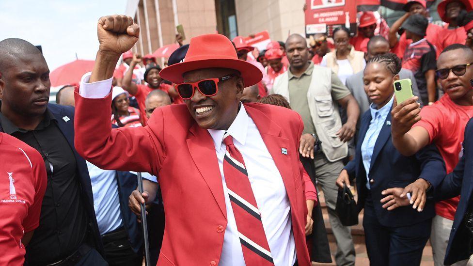 Mokgweetsi Masisi dressed in a red suit and wearing a red hat and sunglasses holds his fist up as he is surrounded by supporters.