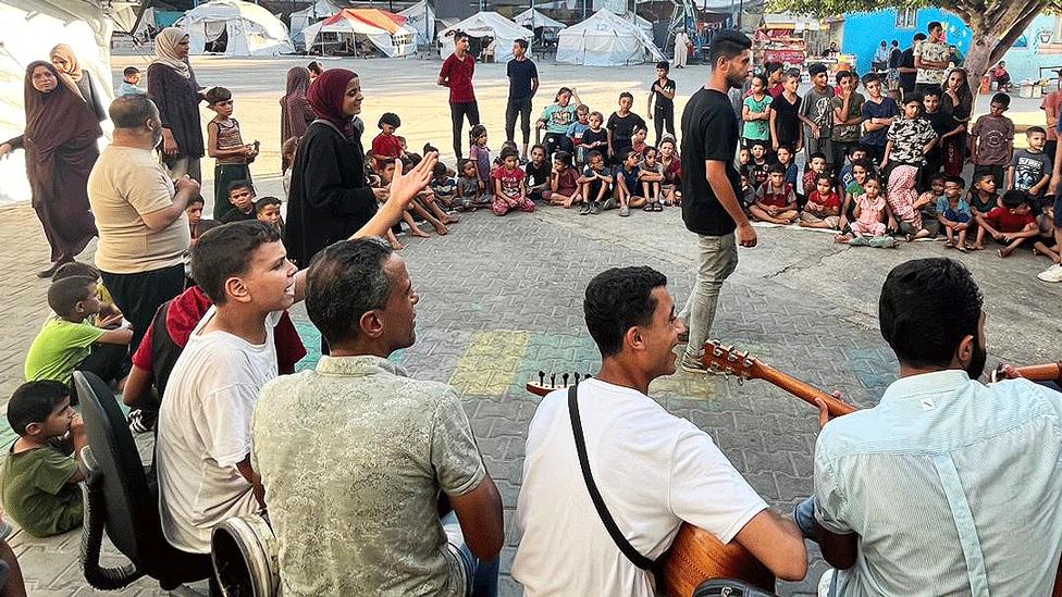 A music session held outside in a Gaza camp led by teachers from the Gaza branch of Palestine’s National Music Conservatory - one man stands, surrounded by several people of all ages sitting down. Two are playing ouds, or guitars. The group is smiling and laughing. 