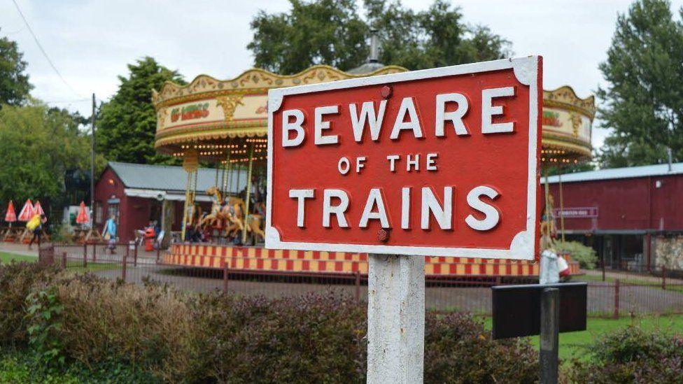 A sign saying 'beware of the trains' with a carousel behind. The bottom of the carousel is red and white checked pattern, and multiple horses and carriages are going round in a circle. 