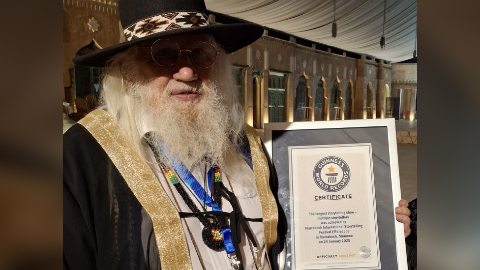 John Row holding his Guiness World Record certificate for longest storytelling show. John has long white hair and a long white beard. He is wearing a black hat, a black jacket with gold details and a blue lanyard. He is also wearing glasses. He is looking at the camera and smiling. 