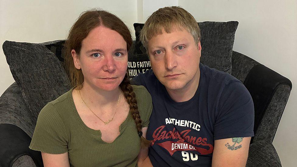 A woman and a man sit side by side on a grey sofa. Th woman has brown hair in a plait and is wearing a green T-shirt. The man has short, light-coloured hair and is wearing a blue T-shirt.