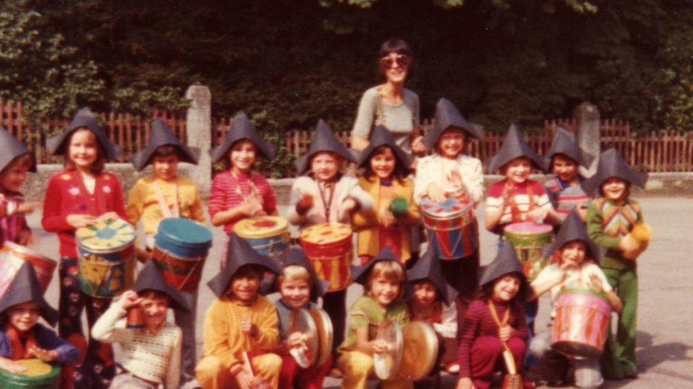 Children wearing black peaked hats kneeling in front of a teacher