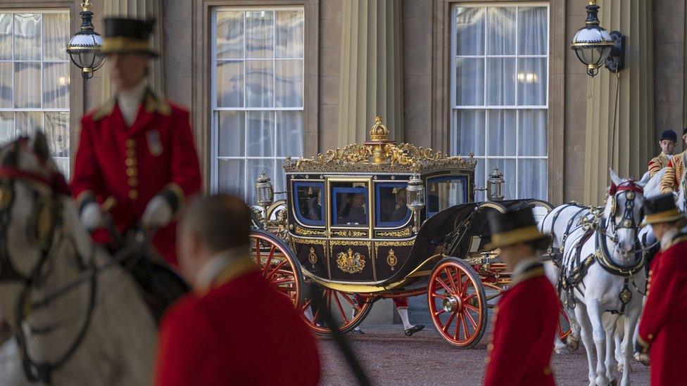 The royal carriage, black and gold, embarks on its journey pulled by white horses. In the foreground are people in red uniforms on horseback.