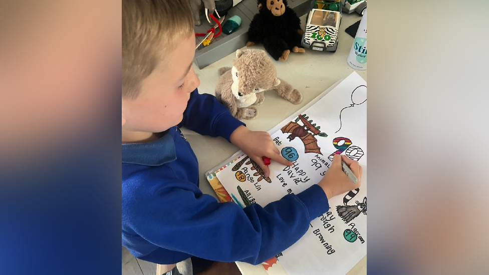A boy in a blue school uniform is drawing pictures of animals on a piece of paper, he is writing Happy 99th birthday to Sir David Attenborough.