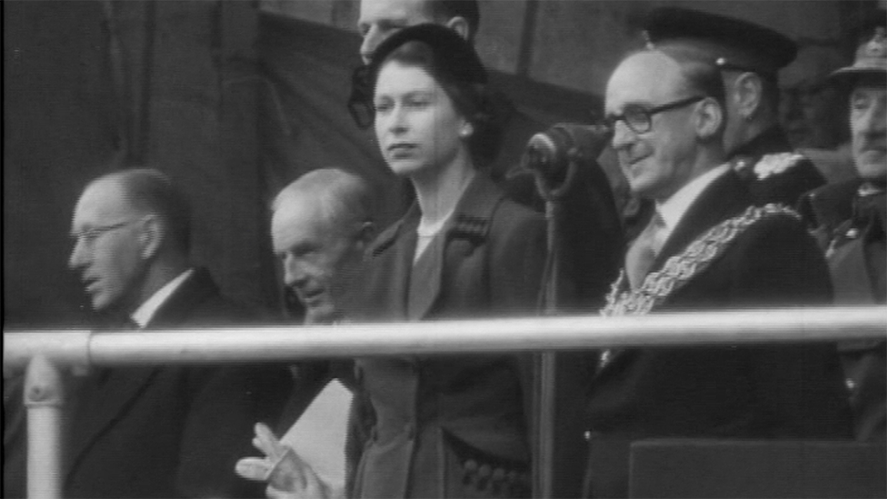 Queen Elizabeth II in 1952 at her first engagement in Wales at the Claerwen dam. The black and white image shows her standing in a suit next to dignitaries with her arms by her side, watching the opening of the reservoir.