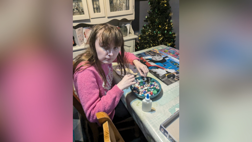 A teenager is sitting at a kitchen table doing arts and crafts. She is wearing a pink top and has long brown hair. There is a bowl of coloured rocks and glue next to her. A Thomas the Tank Engine mat is lying on the table. A Christmas tree is in the background. 