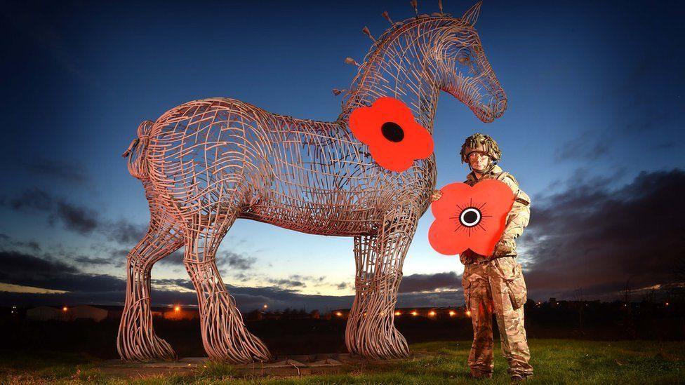 A soldier stands alongside the Heavy Horse sculpture at Baillieston holding the four-petal Scottish poppy 