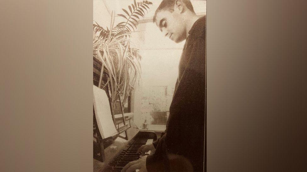 A brown and white sepier style photo of 'Bonehead' aka Paul Arthurs playing the piano in the 1990s when he was part of Oasis. There are plants on top of the piano and he is looking down at the keys. It is the piano going on sale - wooden upright.