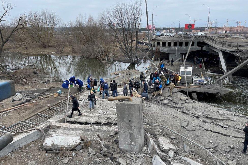 People fleeing Irpin cross the damaged river bridge, which can be seen with the concrete collapsed and girders hanging down into the water