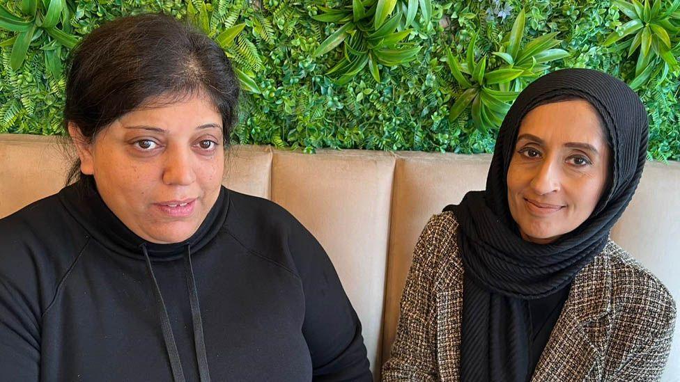 Farzana Jamidar and Raheela Hussain sit together in the cafe. Farzana has her long dark hair tied back and is wearing a black hoodie, Raheela is wearing a brown tweed jacket and black headscarf.