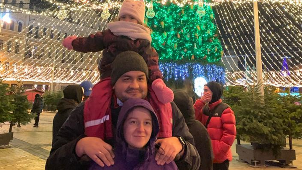 A woman, man and little girl pose in front of a Christmas tree in a square decorated for Christmas