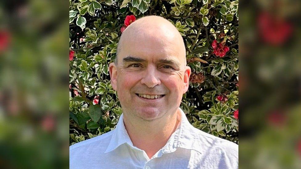 Dr Jones smiling near a bush with flowers. He has brown eyes and is wearing a white shirt.