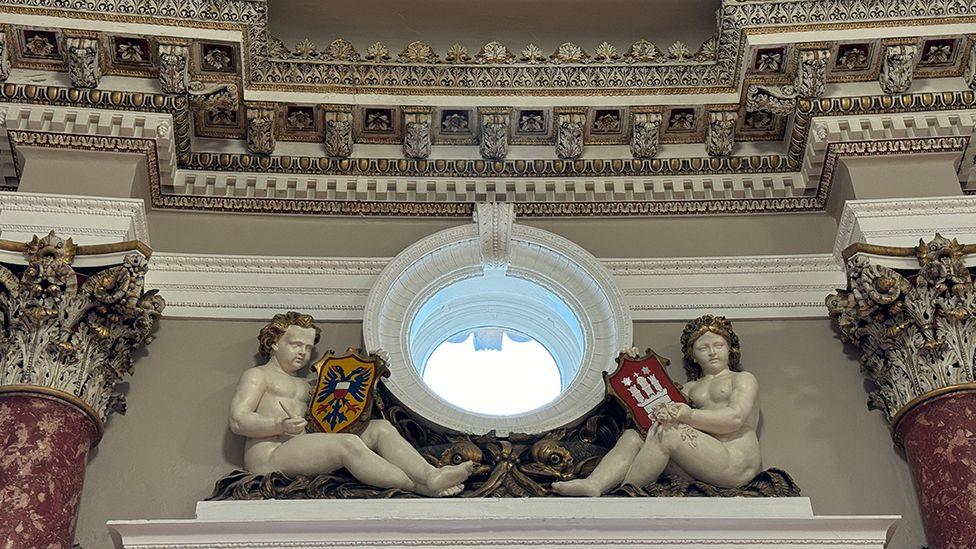 Restored statutes of two white stone figures holding coats of arms sat in front of bronze foliage either side of an oval window 