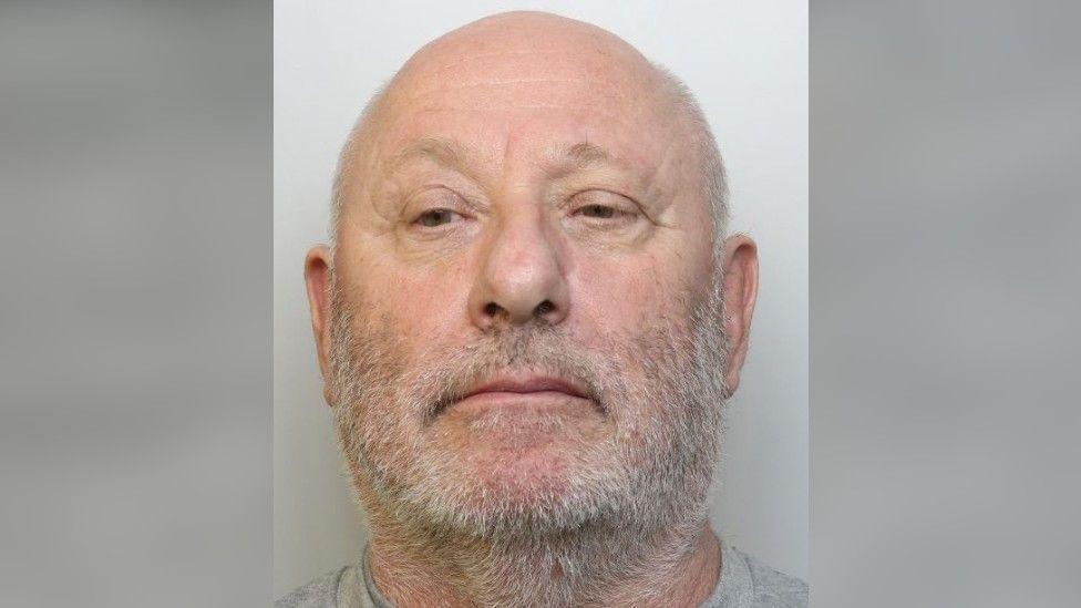 A custody image of Gary Moore. He has very short light grey hair, balding on top, and heavy grey stubble across much of his face. He is wearing a grey t-shirt and is looking slightly away from the camera. 