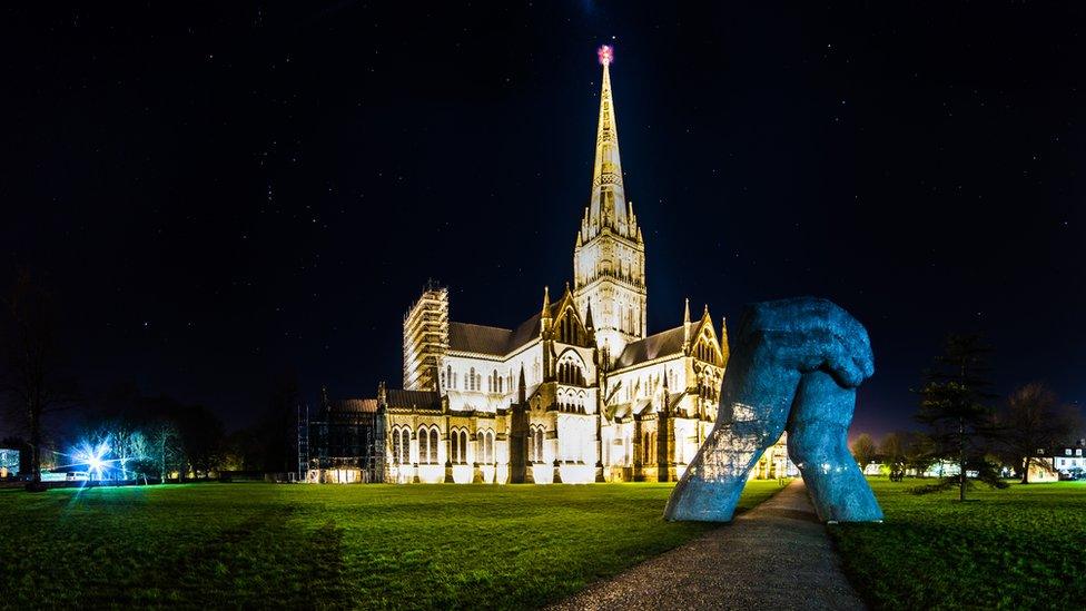 A photo of Salisbury Cathedral at night with a sculpture of hands in front of it