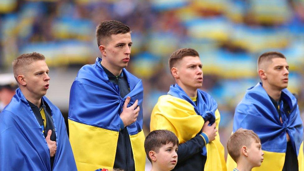 Oleksandr Zinchenko and three of his Ukraine team mates lined up singing their national anthem before a game against England in March. Zinchenko is on the far left and has the Ukraine blue and yellow flag around him. He has short blonde hair and has his hand on his chest. To his right are three other players who are slightly taller than him who all have short brunette hair and all also have a flag draped around them and their hands on their chest.