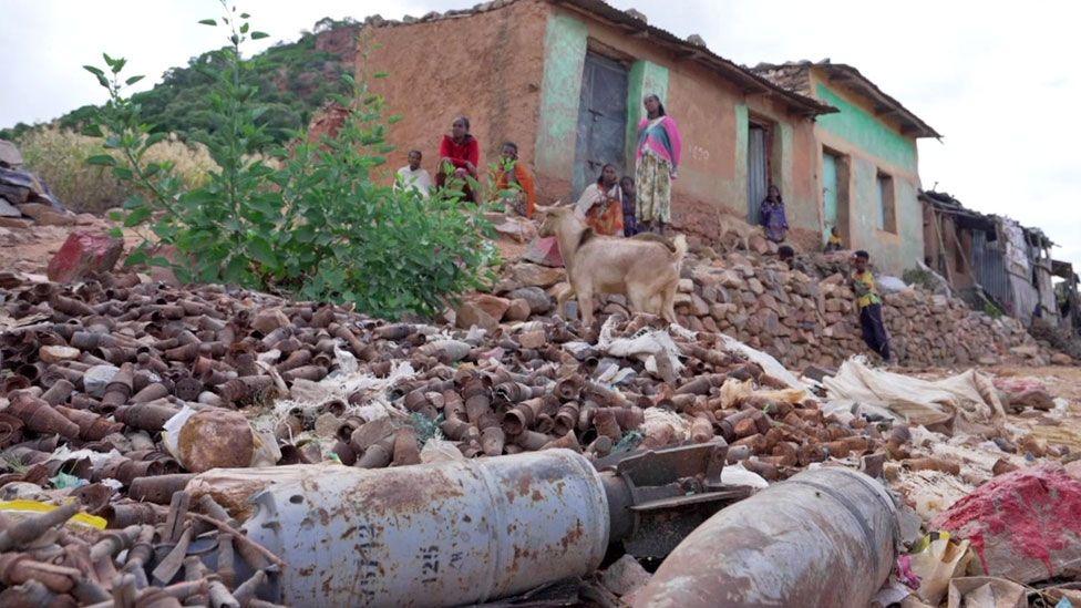 Shells and other rusting ammunition lie on the ground in front of a few houses, some of whose inhabitants are seen sitting outside. A few stones have been painted red as a warning and a goat is nearby. Tigray, Ethiopia - October 2024