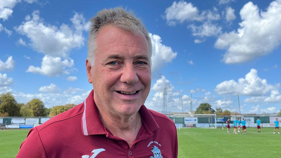 Nigel Arms who has short white hair and is wearing a maroon polo shirt. He is smiling and standing on a pitch, under a blue sky and white clouds.