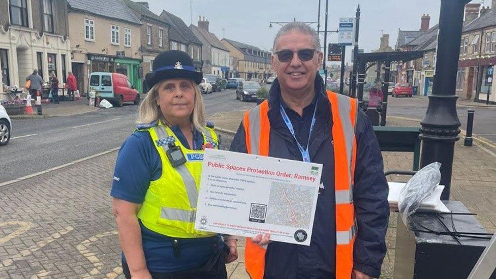 Rachel Martin on the left, she is wearing a police hat over shoulder-length fair hair, and a high-vis police waistcoat over a blue t-shirt. She is standing beside Brian Nevard on the right. He has close-cropped grey hair and is wearing an orange high-vis waistcoat over a blue jacket, while holding a sign that says Public Spaces Protection Order: Ramsey