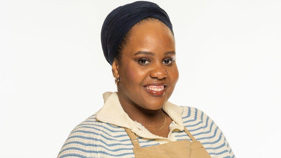 Illyin Morrison wearing a blue and cream striped jumper and a brown baking apron. She is smiling towards the camera.