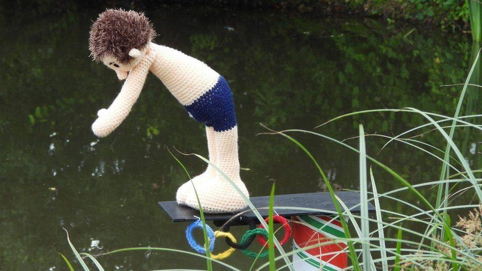 Diver with brown spiky hair leans off a diving board into the pond wearing navy trunks