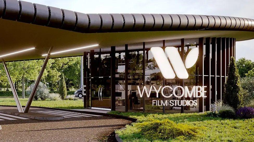 An artist's impression of an entrance building with a sign saying Wycombe Film Studios. The round glass building has a sweeping roof that extends out over a road, held up by a V shape support. It looks very swanky. There are trees behind and lawn with shrubs.