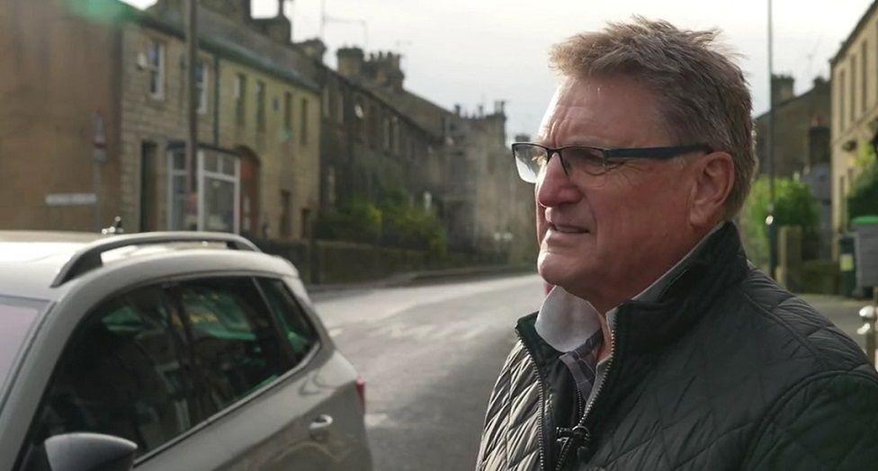 A man with short light-coloured hair, wearing glasses and a shirt and a padded jacket stands on the pavement next to a road. Buildings line both sides of the road behind him. On the left a grey car is passing close to the man.