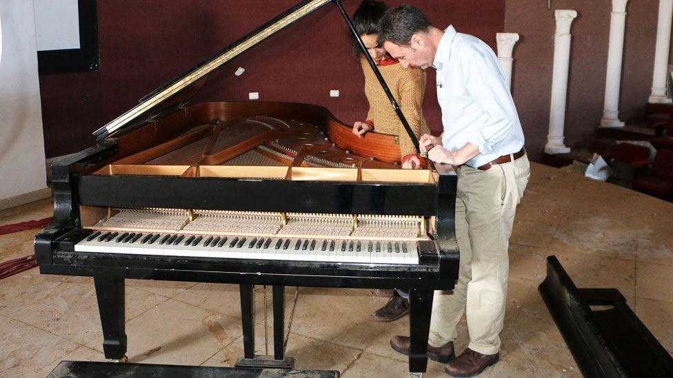 BBC journalist Tim Whewell, dressed in blue shirt and chinos, leans over the hammers of the grand piano - examining it with a French restorer, dressed in a mustard coloured jumper, in 2015