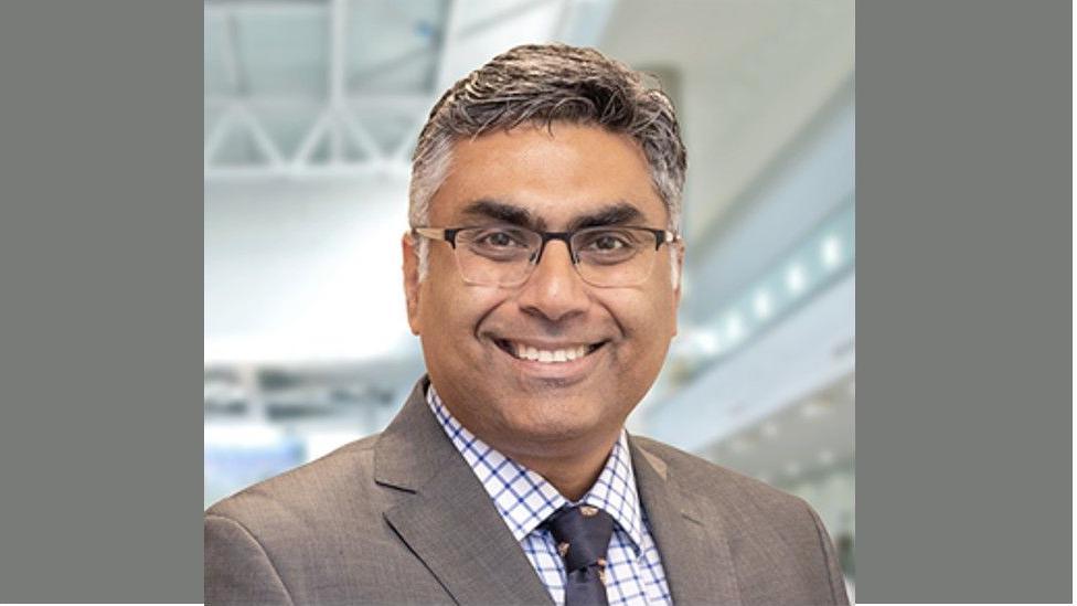 Vikas Khanduja wearing a blazer, blue and white checked shirt, and a blue tie. He also wears square framed glasses and is smiling at the camera. 