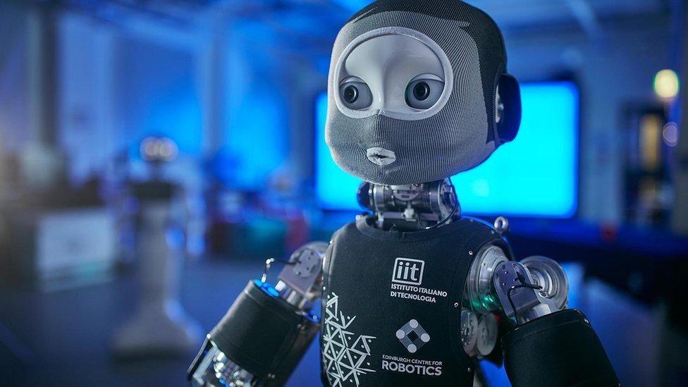 A robot wearing a balaclava, photographed in a lab with a bright blue screen in the background.
