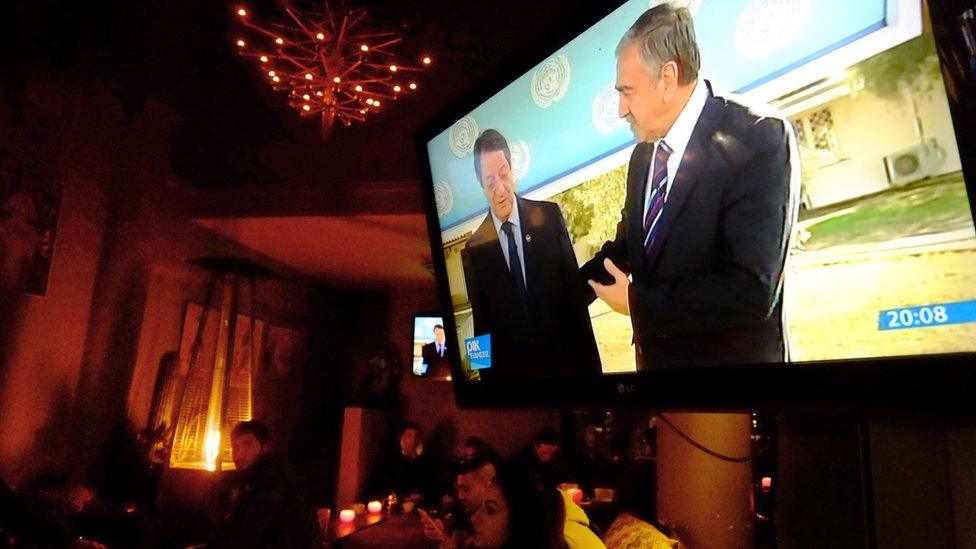 People sit at a coffee bar by a TV showing the Greek Cypriot President Nicos Anastasiades, left. and the leader of the breakaway Turkish Cypriots Mustafa Akinci as they deliver holiday wishes together